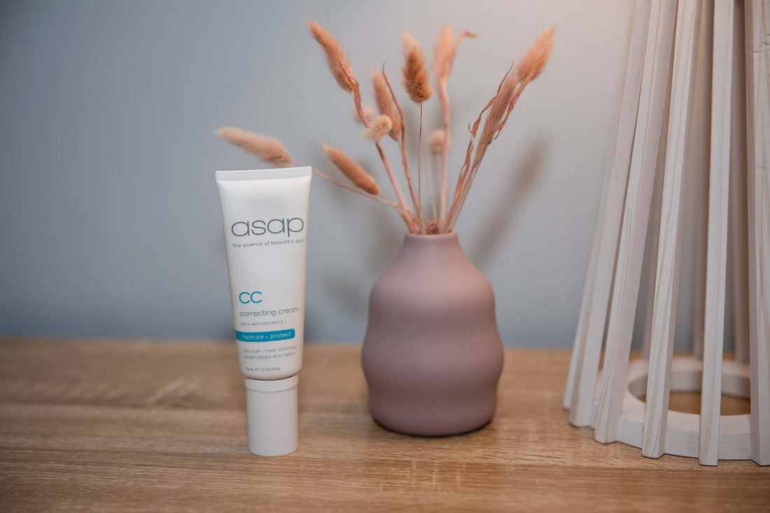 asap correcting cream standing on a wooden table with pink vase full of bunny tail flowers and a lamp to the right 